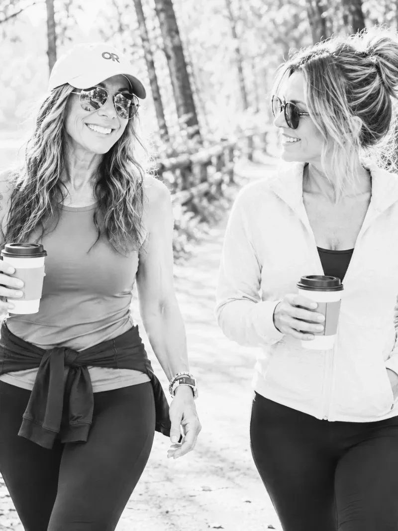 Lisa and a friend smiling as they walk a sunlit trail, both holding cups of coffee