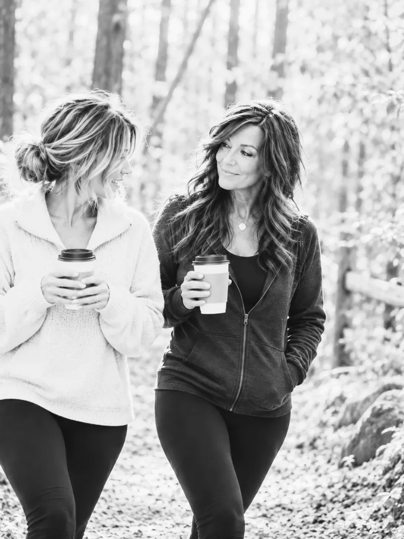 Lisa walking a wooded trail with a friend, both holding cups of coffee