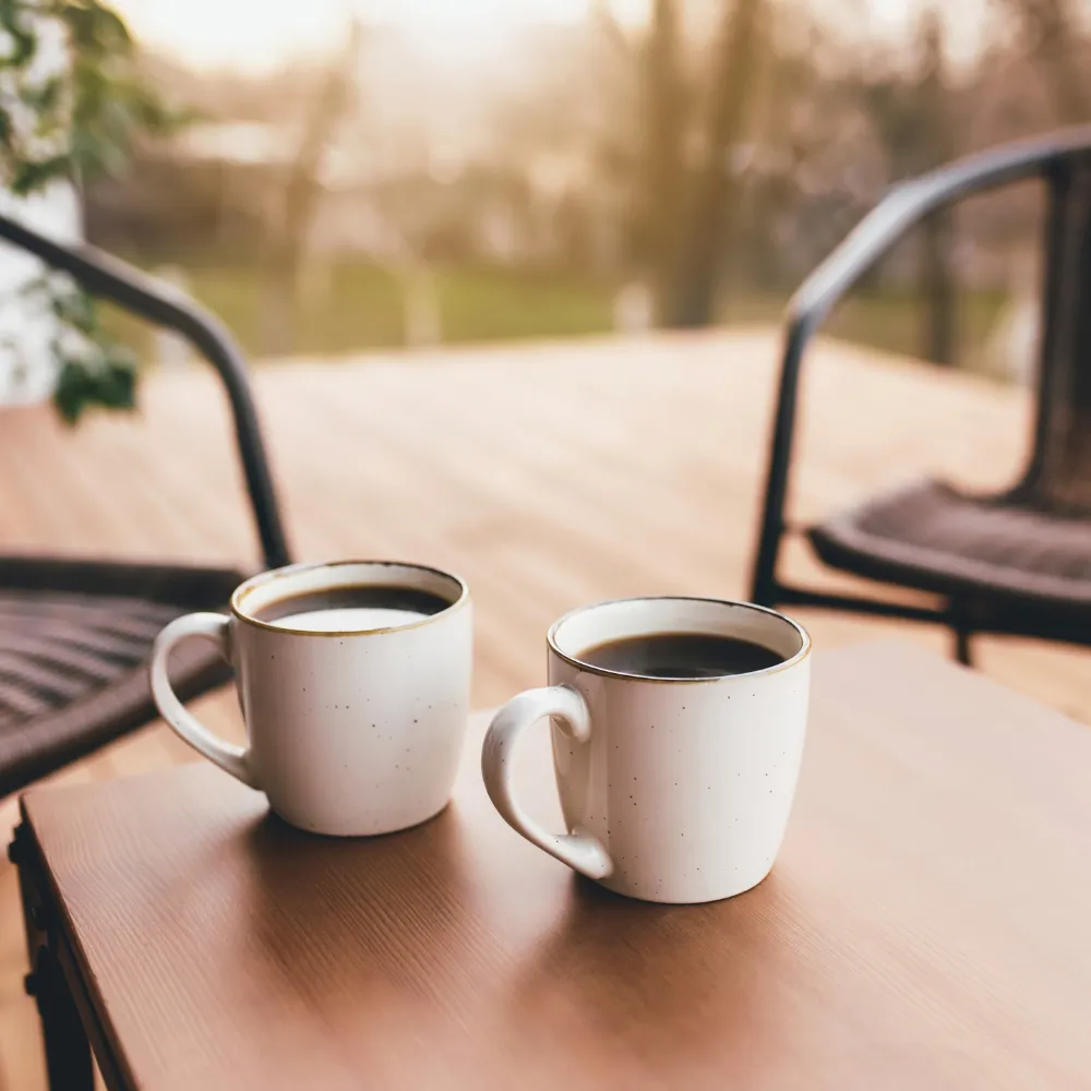 Two mugs of coffee on a wooden table between two chairs on a porch