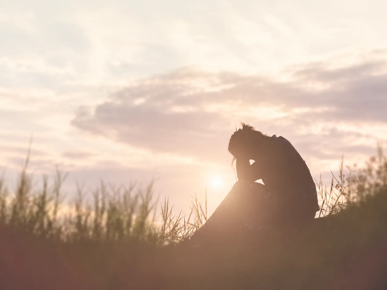Woman sitting alone in tall grass at sunset, head in her hands